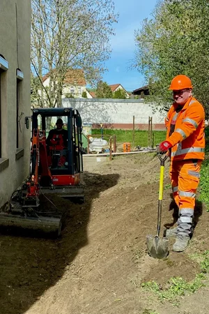 Bild vergr&ouml;&szlig;ern: Auf dem Bild ist im vorderen Bereich ein Bauarbeiter zu sehen, welcher eine Schaufel in den Händen hält. Im Hintergrund steht ein Minibagger, welcher von einem darin sitzenden Mitarbeiter bedient wird.